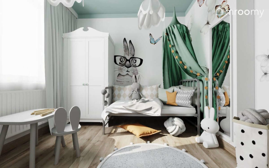 Metalowe łóżko z zielonym baldachimem stolik z krzesełkiem biała szafa i tapeta z królikiem w pokoju małej dziewczynki