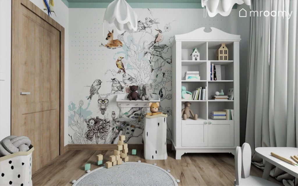 Biała komoda na tle tapety z bajkowym drzewem i zwierzątkami i miętowym sufitem w pokoju małej przedszkolnej dziewczynki