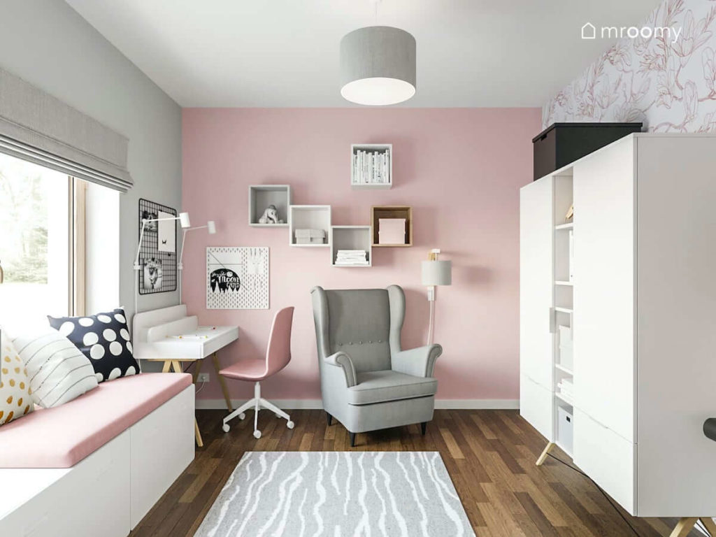 Pokój nastoletniej dziewczynki z szarym fotelem białym biurkiem różową ścianą białą szafą i siedziskiem pod oknem