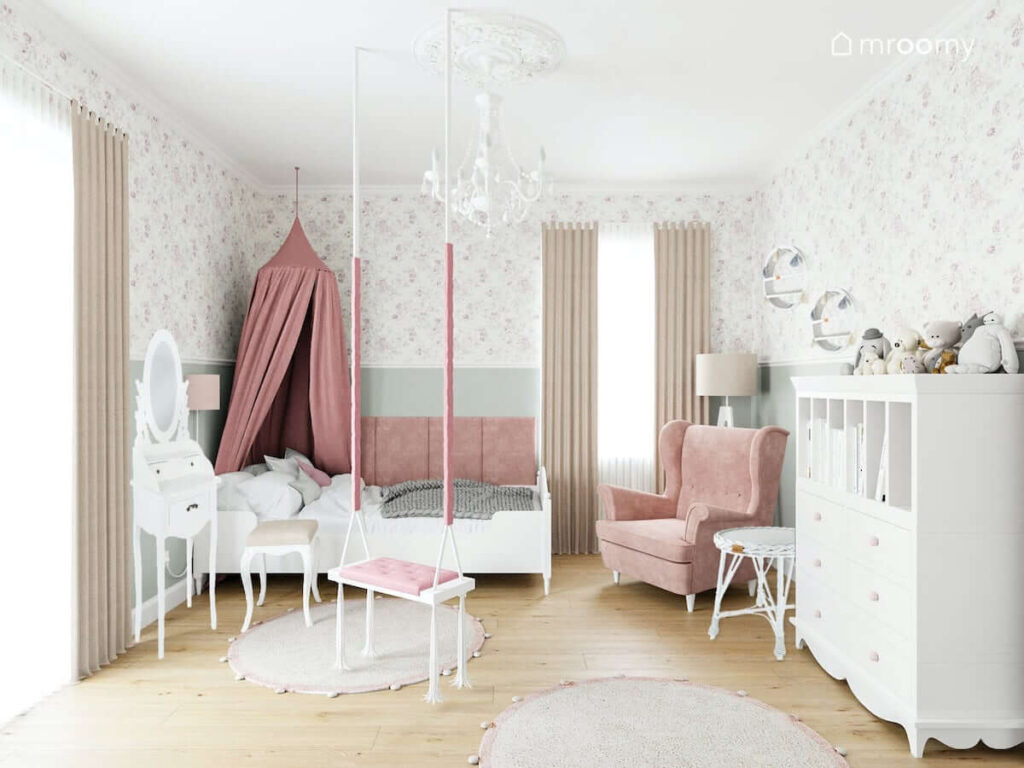 Łóżko ozdobione różowym baldachimem toaletka fotel oraz moduł z szufladami i przegródkami w jasnym pokoju małej dziewczynki zaś u sufitu ozdobny biały żyrandol i huśtawka