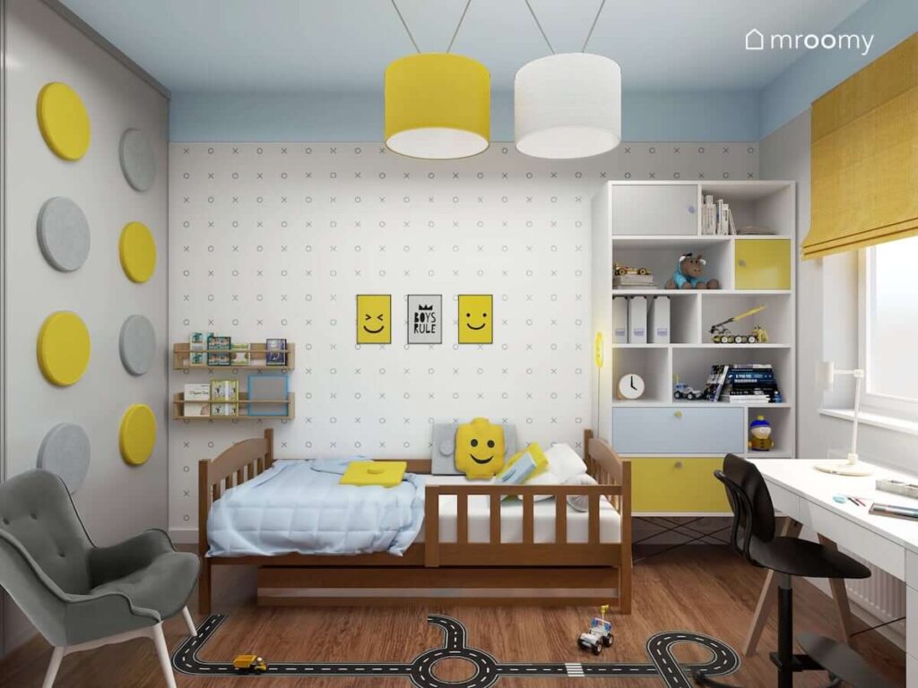 Biało niebiesko żółty pokój dla chłopca z dodatkami w motywach Lego oraz tapetą w kółka i krzyżyki a także pojemnym regałem i naklejką podłogową w kształcie jezdni do tego miękkie panele ścienne w kształcie kolorowych kół