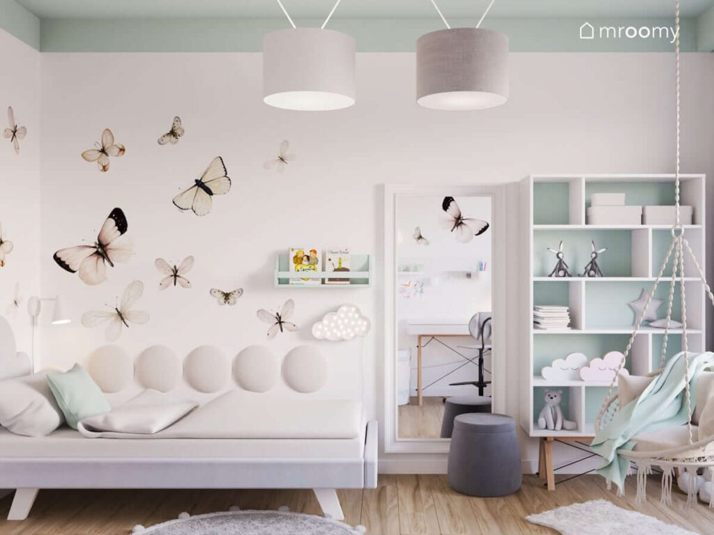 Jasny pokój dla dziewczynki ze ścianami ozdobionymi naklejkami w motyle oraz łóżkiem dużym lustrem regałem i fotelem wiszącym oraz kolorowym sufitem