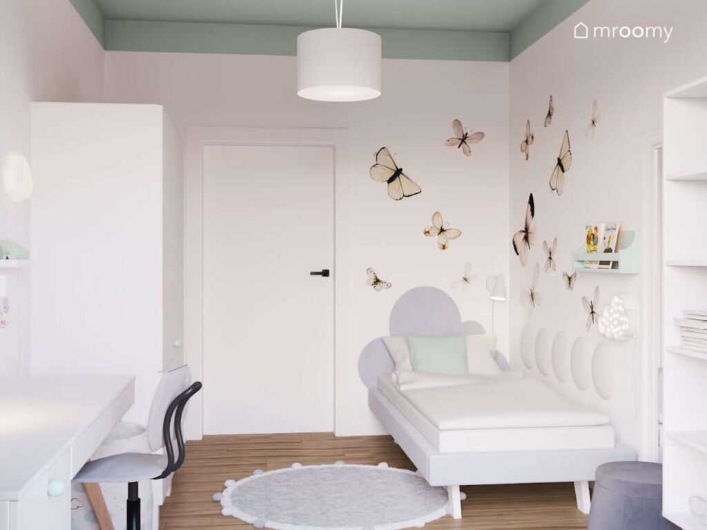 Ściana ozdobiona naklejkami w motyle a pod nią łóżko z zagłówkiem w kształcie chmurki a także biała szafa i biurko w pokoju dla dziewczynki