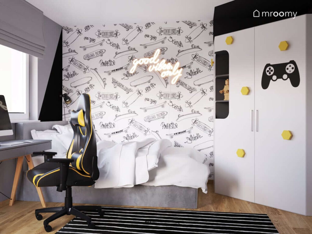 Biało czarno szary pokój dla chłopca miłośnika gier wideo i skateboardingu a w nim szare łóżko biurko z krzesłem gamingowym oraz szafa z żółtymi uchwytami i tapeta w deskorolki