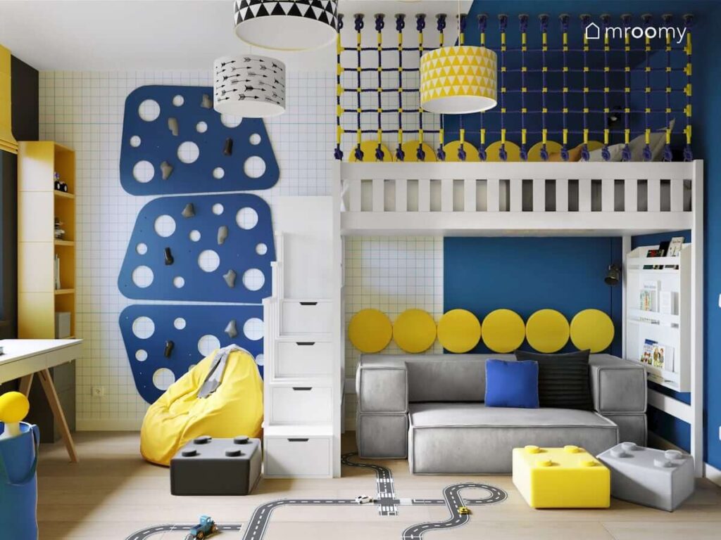 Ścianka wspinaczkowa oraz łóżko na antresoli a pod nim wygodna szara kanapa a całość uzupełniona okrągłymi żółtymi panelami ściennymi w pokoju dla kilkuletniego chłopca