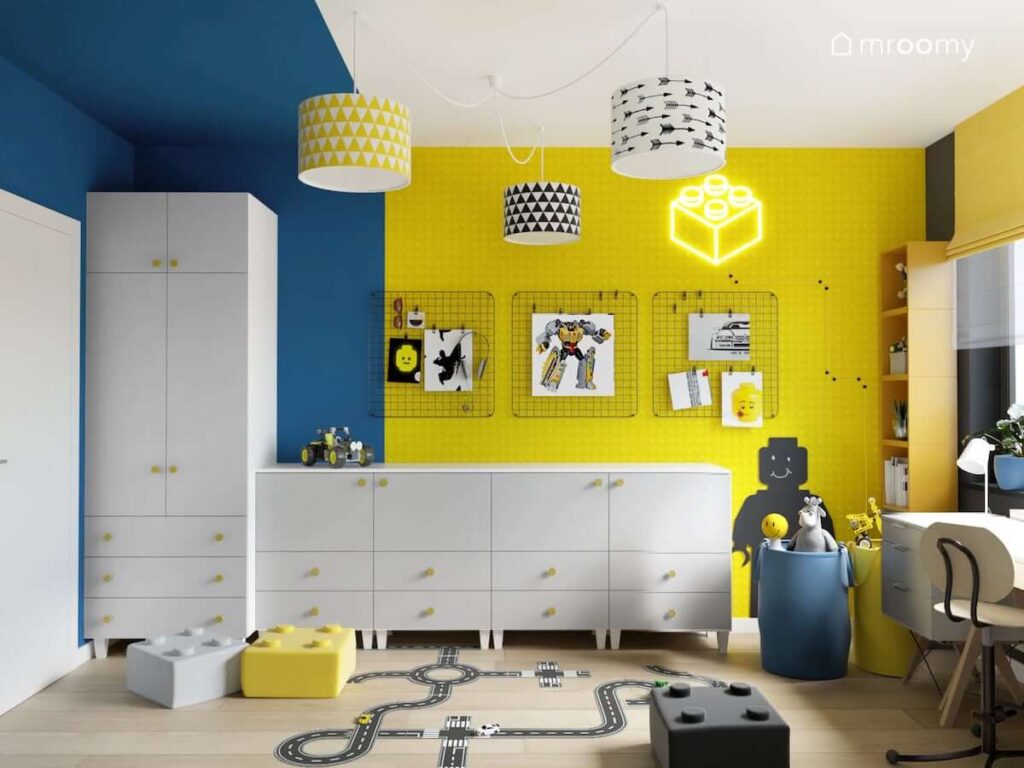 Biało niebiesko żółty pokój dla chłopca w wieku szkolnym z białą szafą modułową a także czarnymi organizerami ściennymi i lampą sufitową z ozdobnymi abażurami