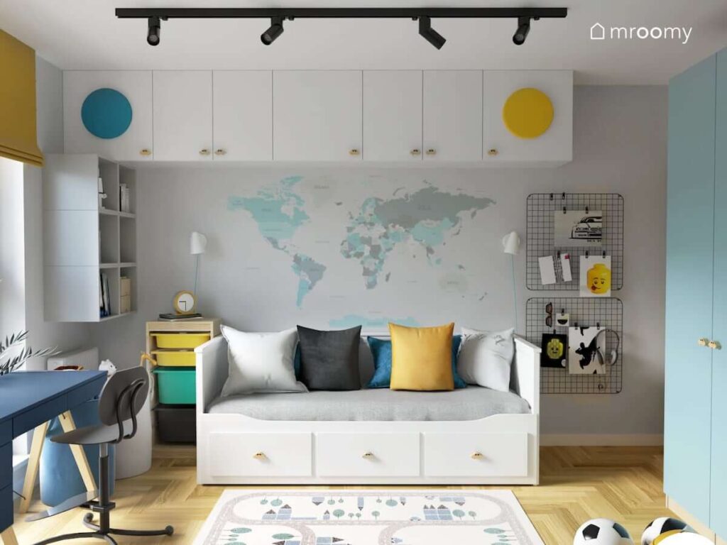 Białe łóżko leżanka z trzema szufladami regał z kolorowymi pojemnikami oraz modułowe szafki a na ścianie organizery ścienne i naklejka w kształcie mapy świata w pokoju dla chłopca