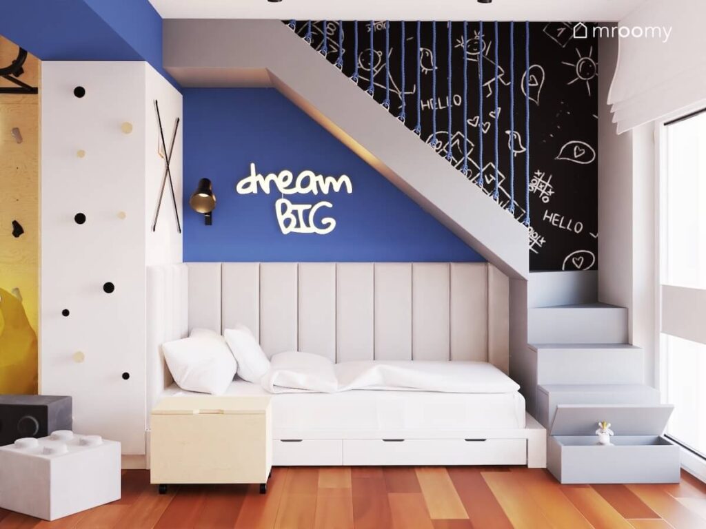 Białe łóżko z szufladami a nad nim panele ścienne i ledon w kształcie napisu oraz ściana pomalowana farbą tablicową w pokoju dwóch chłopców