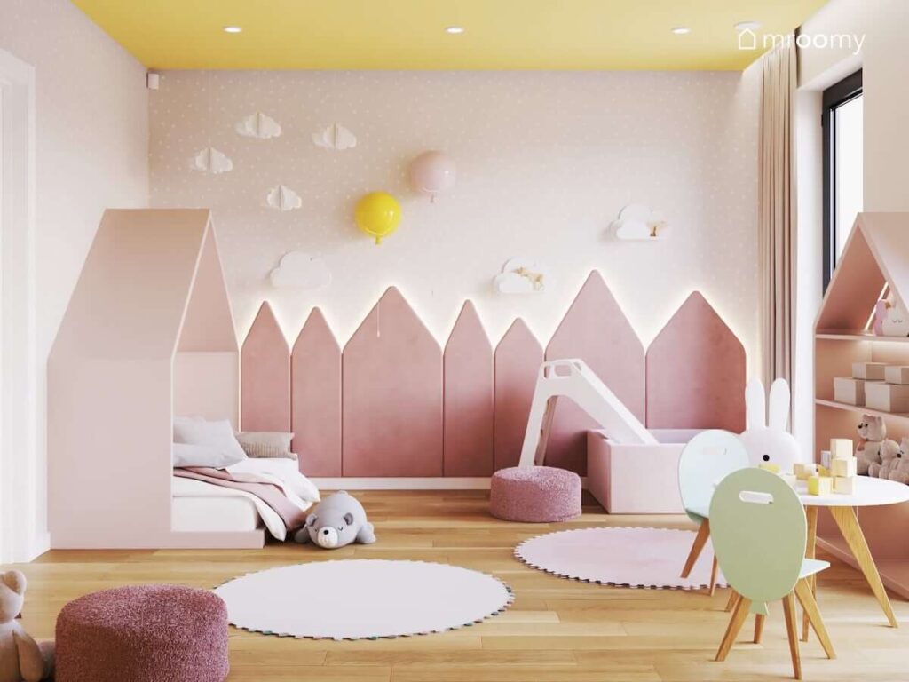 Różowy pokój dla małej dziewczynki z żółtym sufitem panelami ściennymi w kształcie trójkątnych domków oraz ozdobami w kształcie chmurek i kinkietami balonami