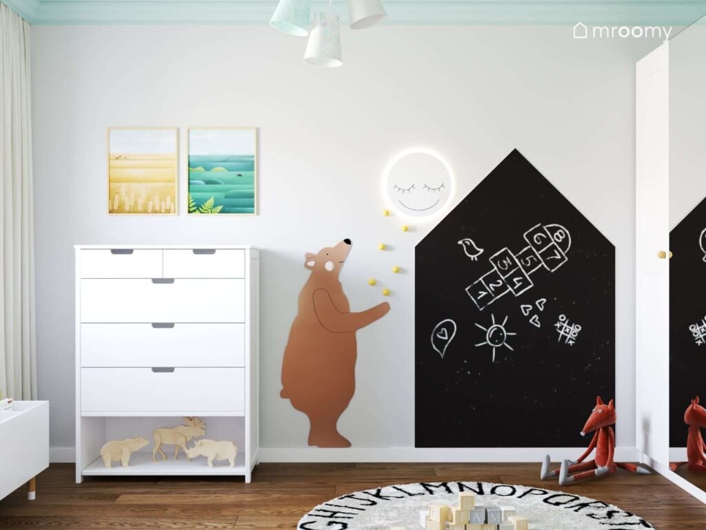 Biała komoda a nad nią ozdobne plakaty z widokami a obok drewniany niedźwiedź lampka w kształcie księżyca i tablica kredowa w kształcie domku w pokoju dla małej dziewczynki