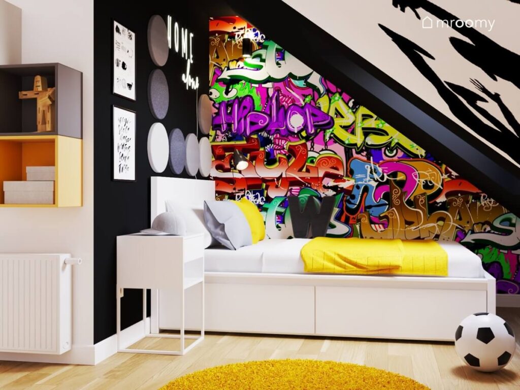 Strefa spania w pokoju dla dziesięciolatka z białym łóżkiem z żółtą narzutą oraz ścianą pokrytą tapetą w graffiti i ścianką ozdobioną szarymi panelami ściennymi oraz ledonem w kształcie napisu