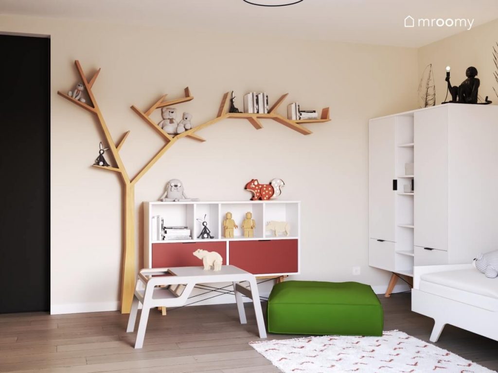 Oryginalna półka w kształcie drzewa oraz biały regał z czerwonymi szufladami zielona pufa sako i biały stolik w jasnym naturalnym pokoju dziewczynki
