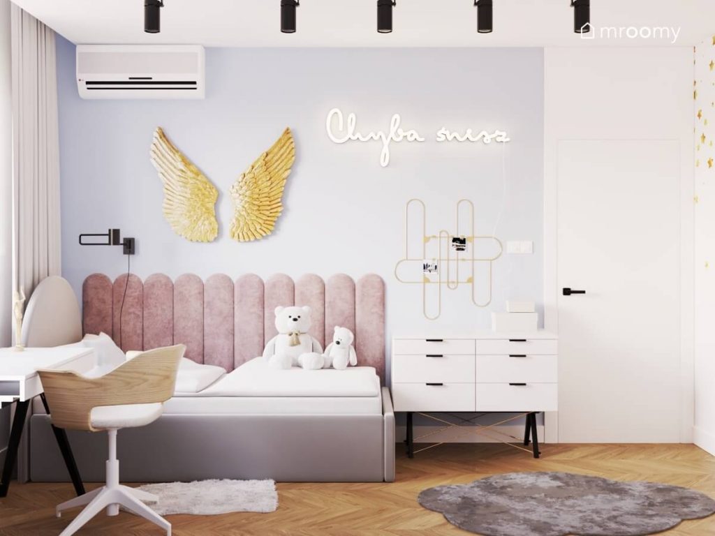 Szare łóżko w pokoju dziewczynki uzupełnione różowymi panelami ściennymi a nad nim złote anielskie skrzydła i ledon w kształcie napisu a obok biała komoda i złota tablica na notatki