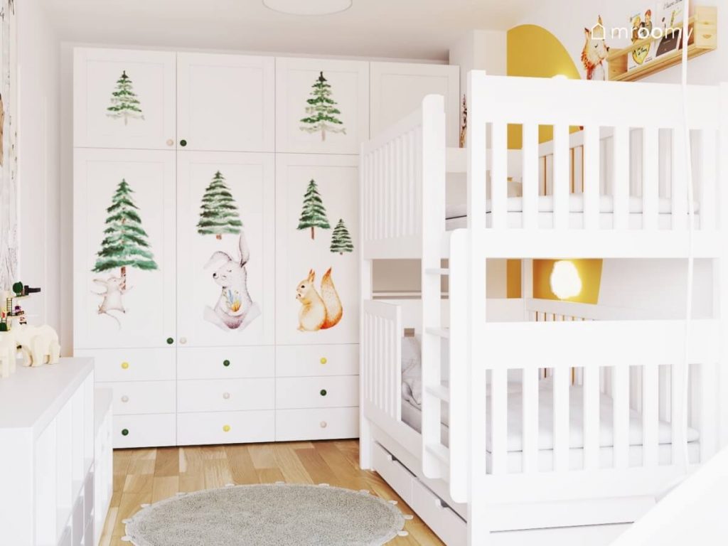 Duża biała szafa uzupełniona naklejkami w kształcie choinek i zwierząt leśnych oraz kolorowymi gałkami oraz białe łóżko piętrowe w pokoju dla małego rodzeństwa