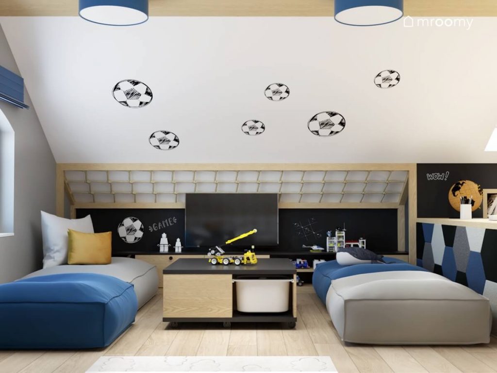 Strefa rozrywki w poddaszowym pokoju dla chłopca w wieku szkolnym a w niej telewizor w zaaranżowanej bramce stolik do zabawy oraz niebieskie i szare pufy