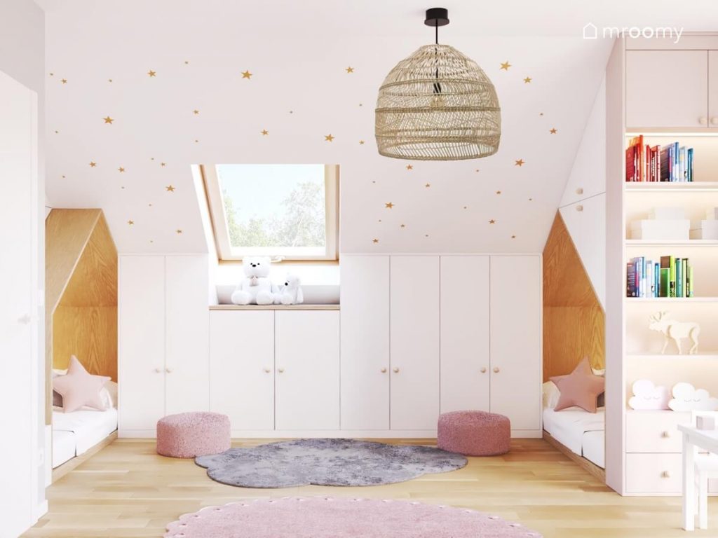 Dwa łóżka domki a także zabudowa meblowa na ściance kolankowej a na podłodze dwie różowe pufy oraz szary i różowy dywan a na suficie lampa w bambusowym kloszu w poddaszowym pokoju małych sióstr
