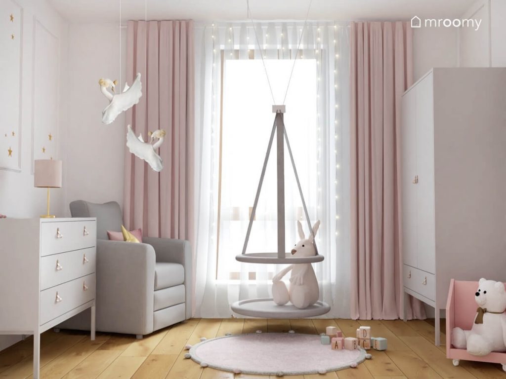 Biały pokój dla dziewczynki z jasnymi meblami szarym fotelem wiszącą huśtawką kojcem ozdobami w kształcie łabędzie różowymi zasłonami oraz szarym dywanem