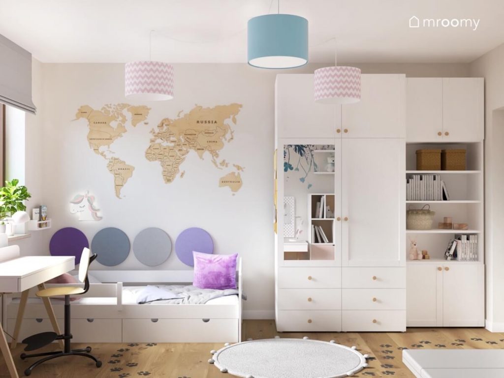 Białe łóżko z szufladami uzupełnione panelami ściennymi w różnych odcieniach fioletowego a nad nim mapa świata ze sklejki a obok biała szafa z lustrzanym frontem a na suficie lampy w kolorowych abażurach