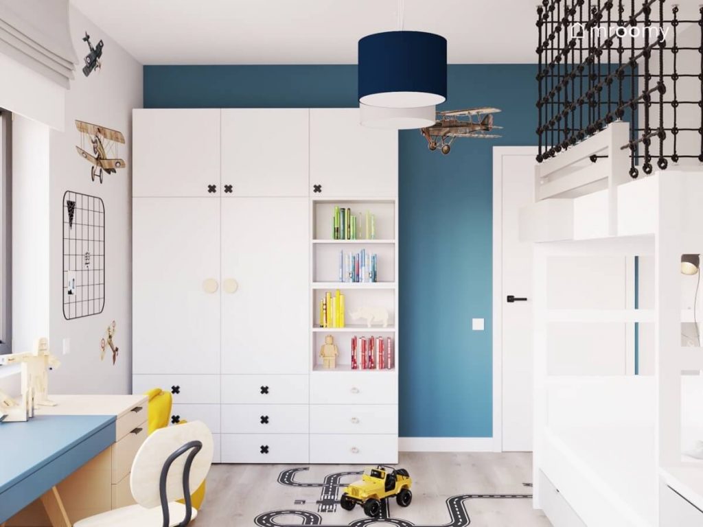 Biała szafa modułowa z drewnianymi i czarnymi gałkami w kształcie iksów a za nią niebieska ściana w pokoju dla chłopca