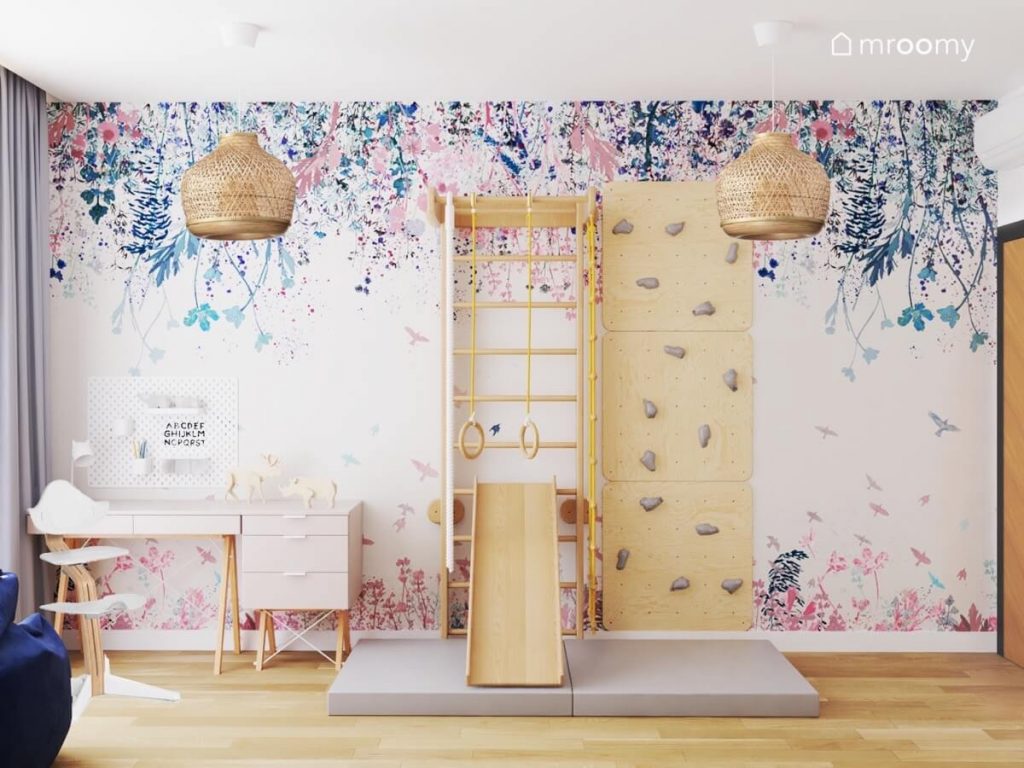 Drewniana drabinka gimnastyczna ze zjeżdżalnią oraz ścianka wspinaczkowa ze sklejki a obok strefa nauki a na ścianie kwiecista tapeta w pokoju dziewczynki