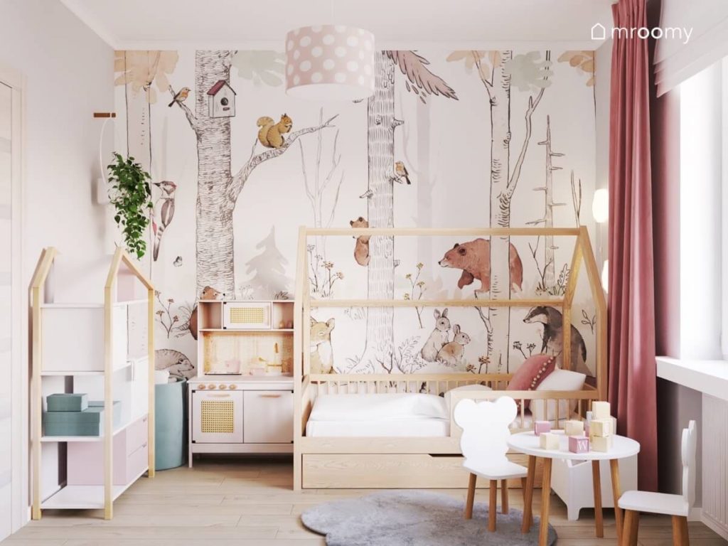Ściana pokryta leśną tapetą a także drewniane łóżko domek kuchenka dla dzieci i stoliczek w pokoju małej dziewczynki