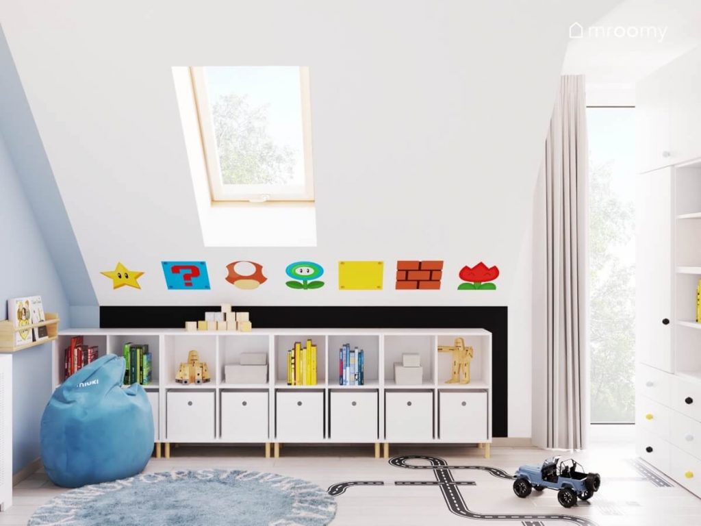 Niskie regały z pojemnikami a nad nimi naklejki z gry Mario a na podłodze niebieska pufa dywan z alfabetem i naklejka w kształcie jezdni w poddaszowym pokoju chłopca