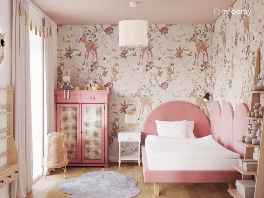 Ściana pokryta tapetą w zwierzęta i kwiaty a także różowa szafa a także różowe tapicerowane łóżko w pokoju kilkulatki