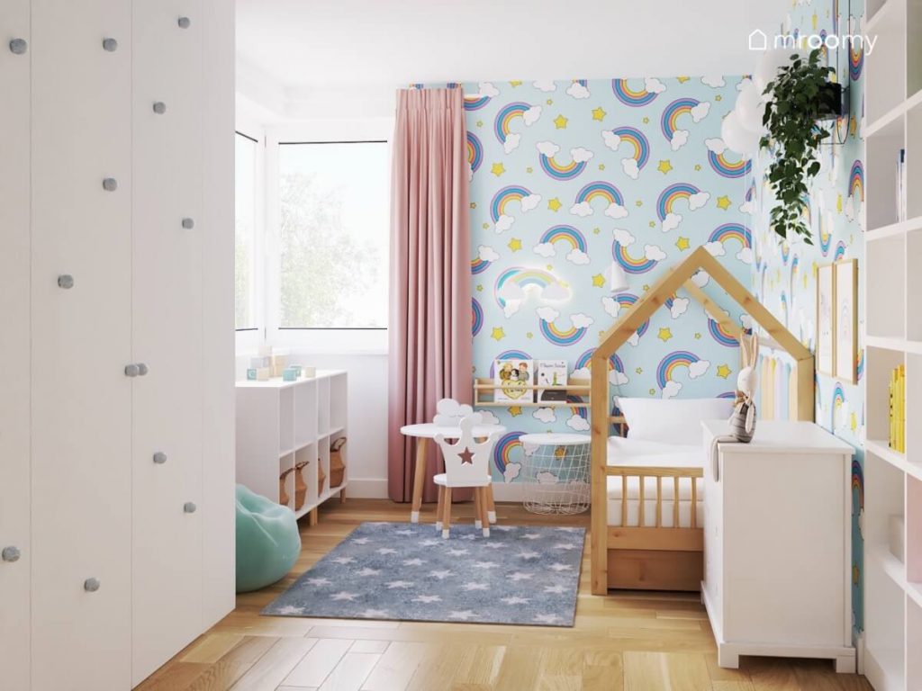 Jasny pokój dziewczynki a w nim biała szafa uzupełniona błękitnymi gałkami drewniane łóżko domek stolik z krzesełkiem z oparciem w kształcie korony a na podłodze dywan w gwiazdki a na ścianie tapeta w tęcze