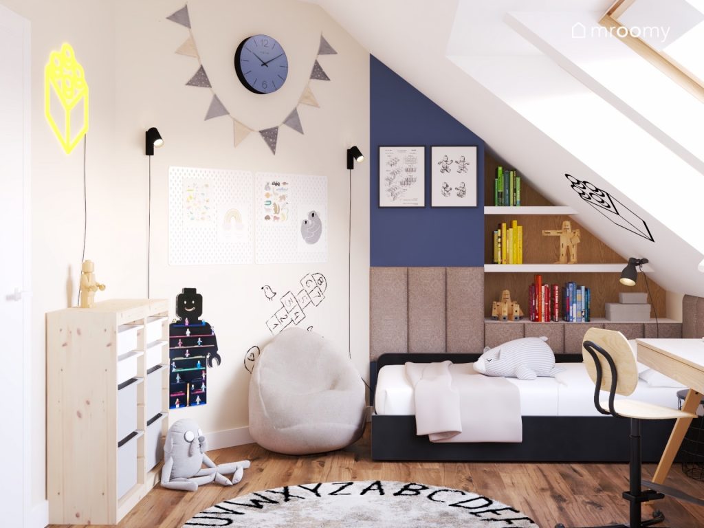 Poddaszowy pokój dla chłopca a w nim kremowo biało niebieskie ściany czarne łózko białe organizery tablica kredowa w kształcie ludzika Lego oraz ciemnoniebieski zegar i girlanda chorągiewek