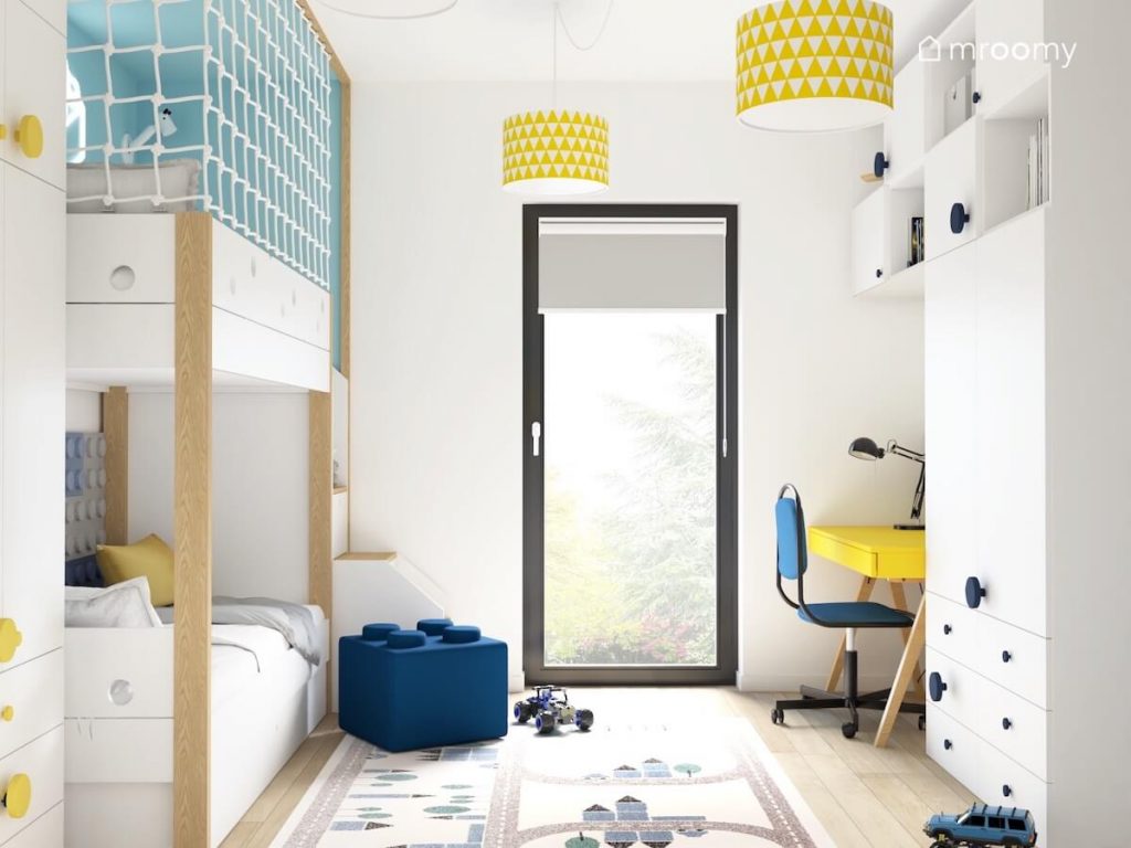 Jasny pokój dla chłopca a w nim biało drewniana antresola pufka w kształcie klocka dywan z miastem oraz białe szafki i żółte biurko