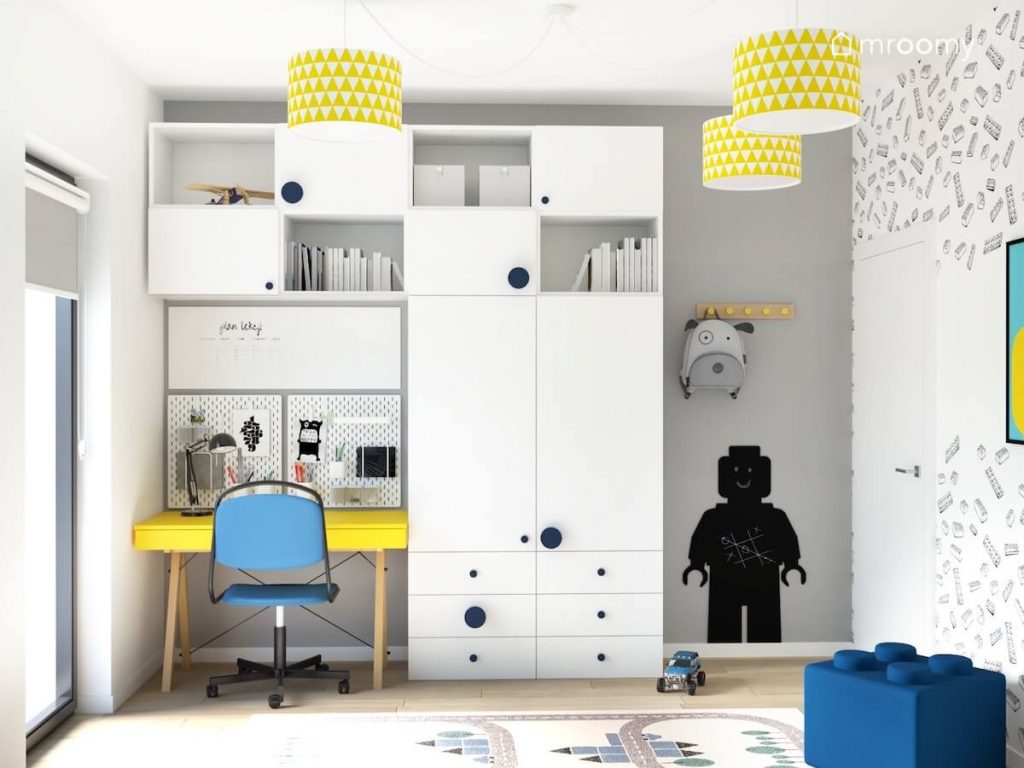 Białe meble modułowe z granatowymi uchwytami oraz żółte biurko z niebieskim krzesłem a obok tablica kredowa w kształcie ludzika Lego w biało szarym pokoju chłopca