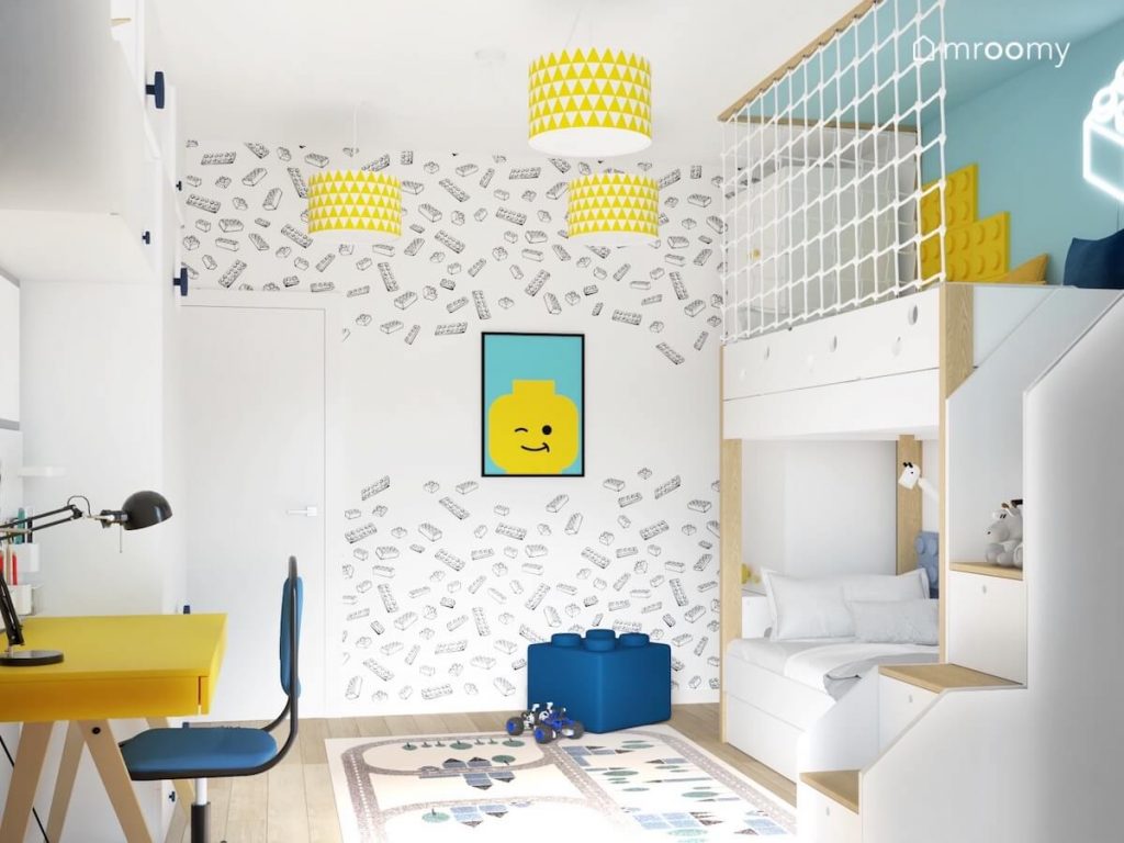 Ściana pokryta tapetą w klocki Lego a na niej plakat również z Lego a na suficie lampy z biało żółtymi abażurami w trójkąty w pokoju dla chłopca
