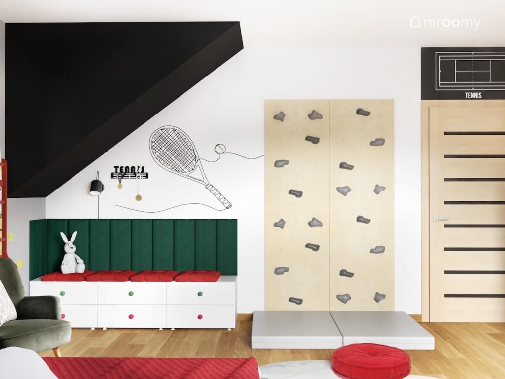 Białe szafki z czerwonymi i zielonymi gałkami a na nich siedzisko z poduszek i paneli ściennych obok ścianka wspinaczkowa a na ścianie tenisowa naklejka w pokoju dziewczynki