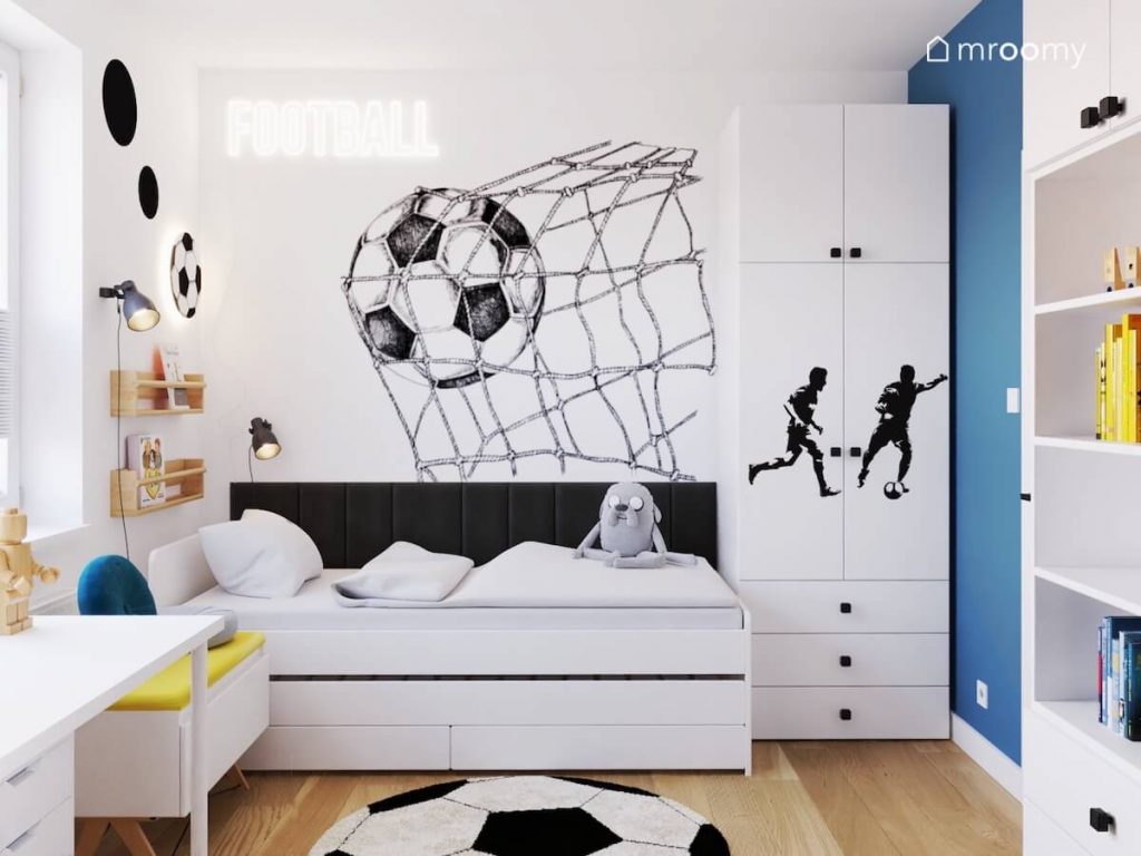 Białe łóżko uzupełnione ciemnymi panelami ściennymi a obok szafa uzupełniona naklejkami z piłkarzami a na ścianie ledon w kształcie napisu oraz naklejka w kształcie piłki wpadającej w bramkę w pokoju chłopca
