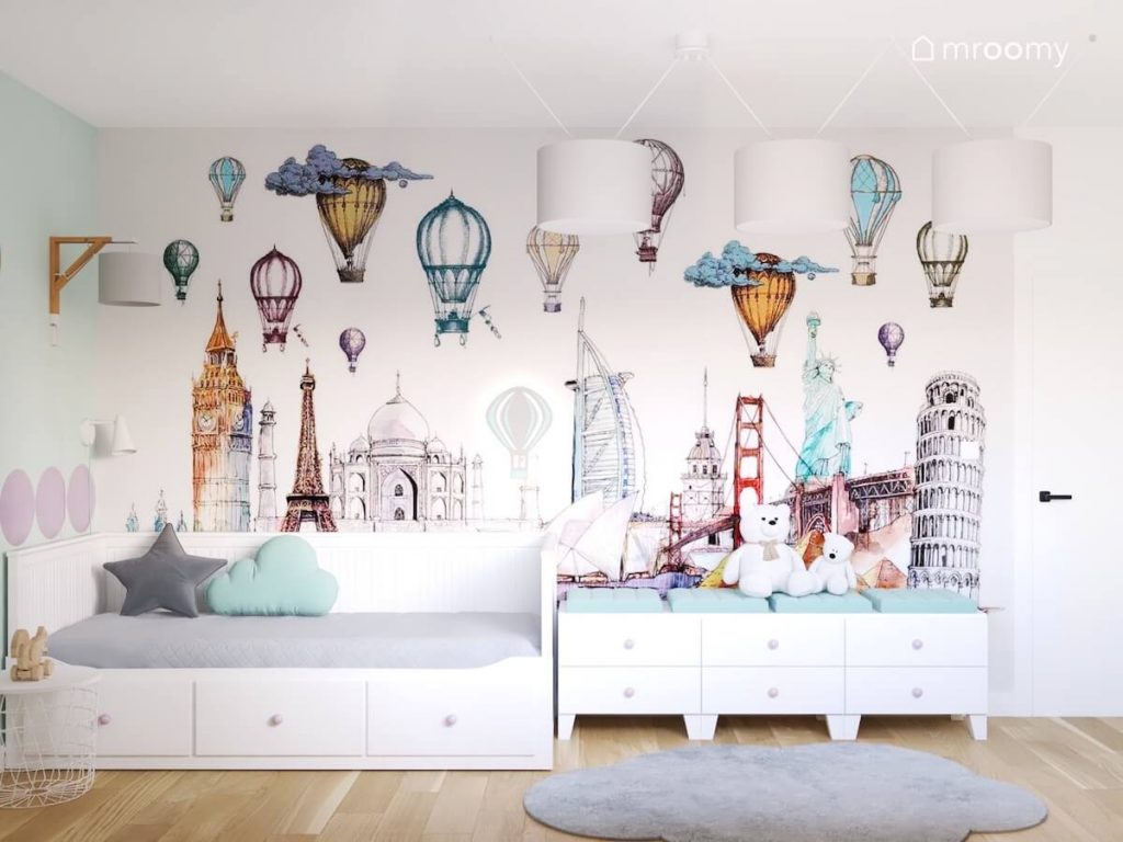 Ściana pokryta tapetą z zabytkami z różnych stron świata oraz balonami a pod nią białe łóżko oraz szafki z miętowymi siedziskami w pokoju dziewczynki
