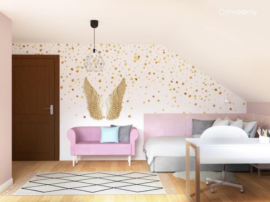 Ściana pokryta tapetą w złote kropki a na niej złote skrzydła a pod nią różowa sofa oraz szare łóżko uzupełnione różowymi panelami w poddaszowym pokoju dziewczynki