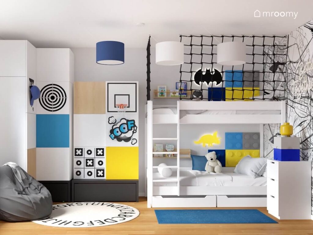 Biało czarna szafa z kolorowymi nakładkami na fronty i koszem do koszykówki a obok białe piętrowe łóżko uzupełnione kolorowymi panelami ściennymi w kształcie klocków w pokoju dwóch chłopców