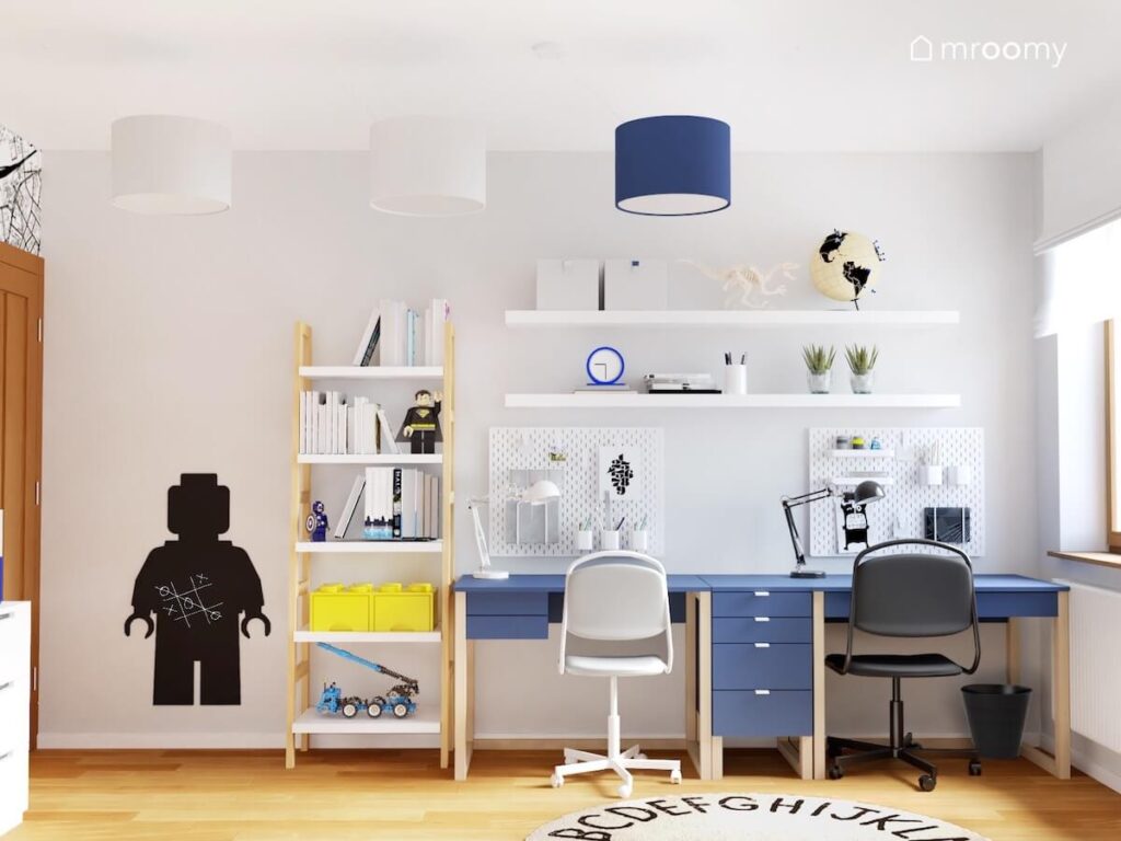 Strefa nauki w pokoju dwóch braci a w niej dwa granatowe biurka ze wspólnym kontenerkiem organizery oraz półki a obok biało drewniany regał i tablica kredowa w kształcie ludzika Lego