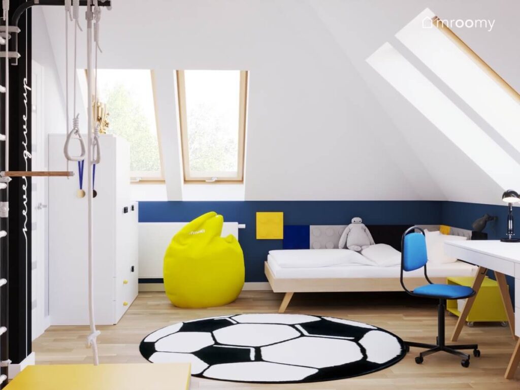 Biało granatowy poddaszowy pokój dla chłopca a w nim białe i drewniane meble żółta pufa i dywan w kształcie piłki