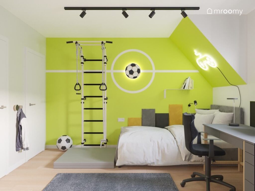 Soczyście zielona ściana w pokoju chłopca a na niej lampka w kształcie piłki oraz drabinka gimnastyczna a pod spodem łóżko