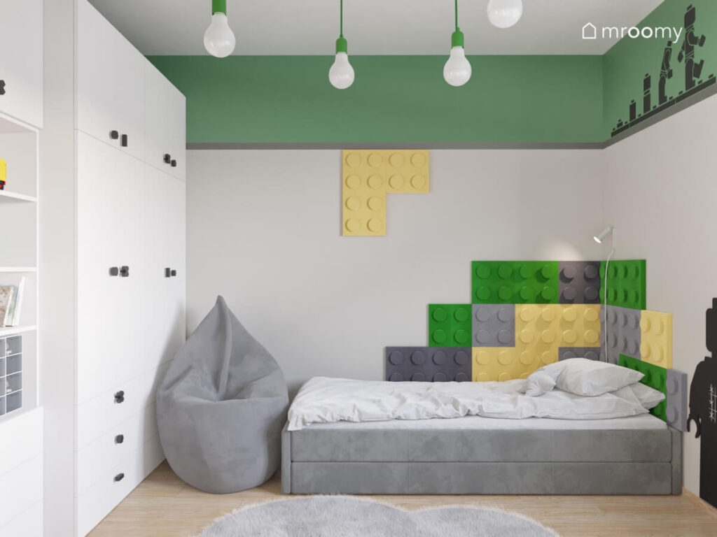 Strefa spania w szaro zielonym pokoju dwóch chłopców a w niej szare tapicerowane łóżko duża szara pufa a na ścianie kolorowe panele w kształcie klocków