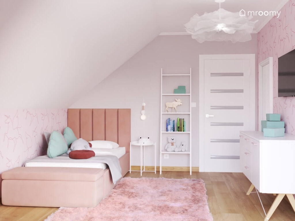 Różowe tapicerowane łóżko biały regał drabinka na podłodze różowy dywan a u sufitu ozdobna lampa w poddaszowym pokoju dziewczynki