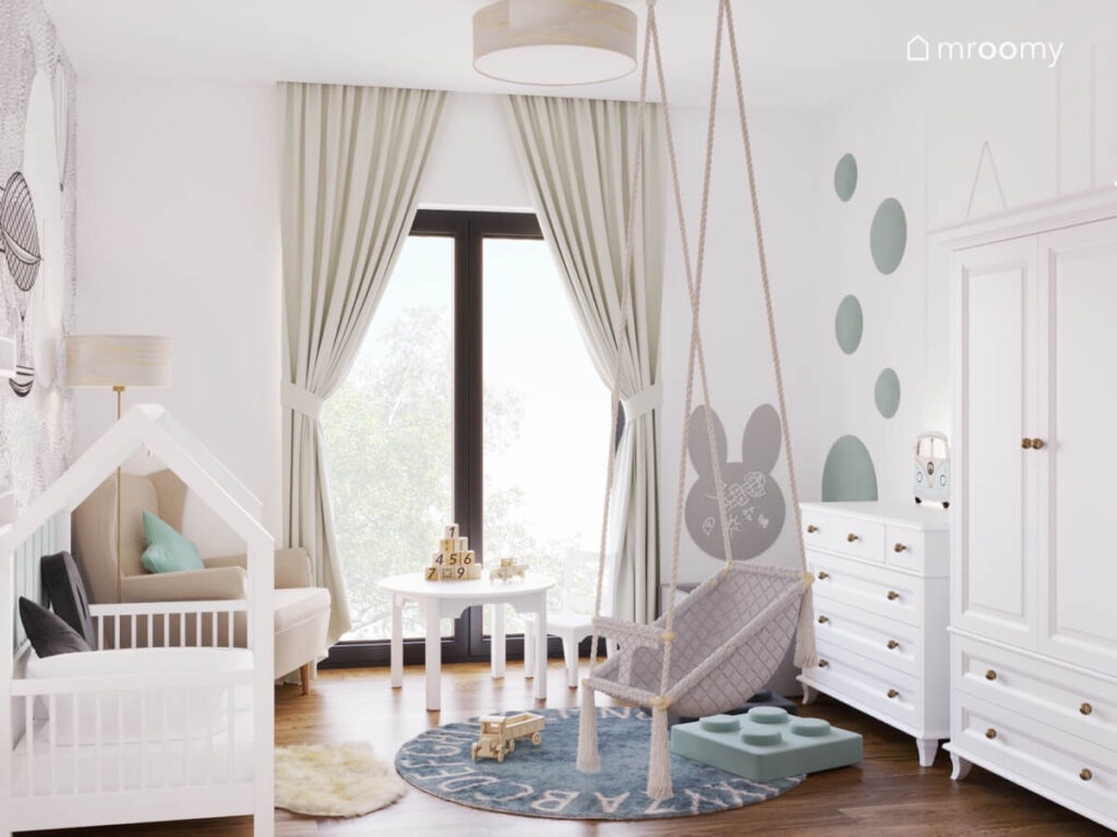 Jasny pokój dla niemowlaka z białymi meblami huśtawką wiszącą oraz jasnoniebieskimi dodatkami