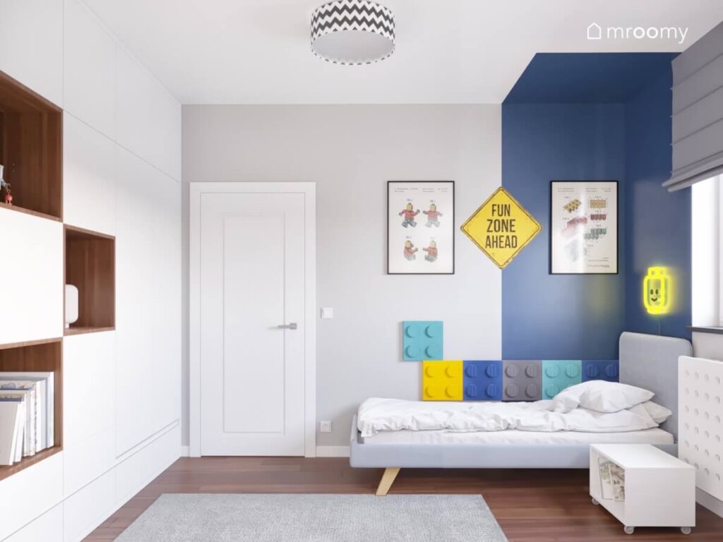 Biało szaro niebieski pokój dla chłopca z szarym tapicerowanym łóżkiem uzupełnionym kolorowymi panelami w kształcie klocków