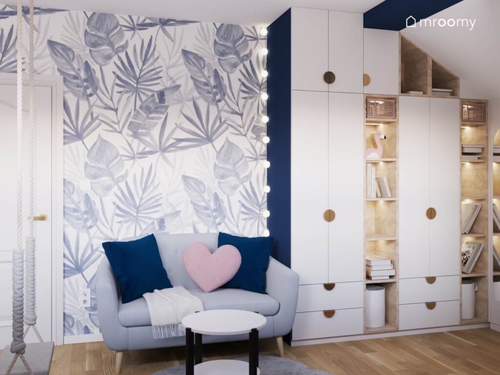 Biała szafa z drewnianymi półkami oraz szara sofa ze stolikiem a na ścianie tapeta w liście w pokoju dla dziewczynki