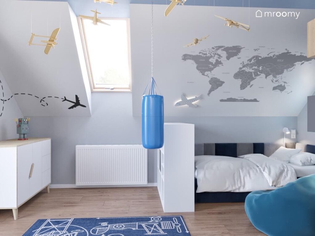 Skos w pokoju chłopca ozdobiony naklejką z mapą świata i samolotem pod nim łóżko a na środku dywan worek treningowy oraz drewniane samolociki