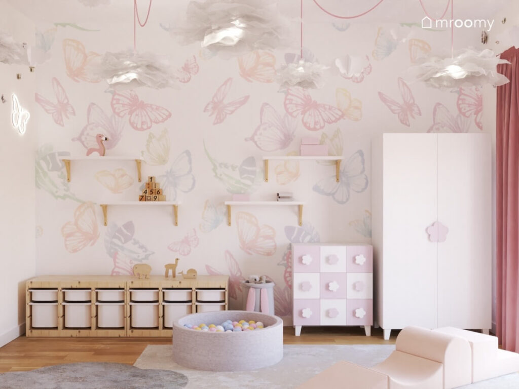 Ściana w pokoju dziewczynki pokryta tapetą w motyle a na niej półki niski regał z pojemnikami oraz meble z uchwytami w kształcie kwiatków